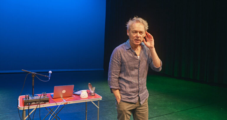 Een man staat op een podium met een laptop en een tafel vol apparatuur, terwijl hij in de richting van het publiek spreekt.