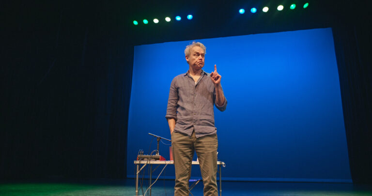 Een man staat op een podium met een blauwe achtergrond en spreekt, met één hand opgeheven.