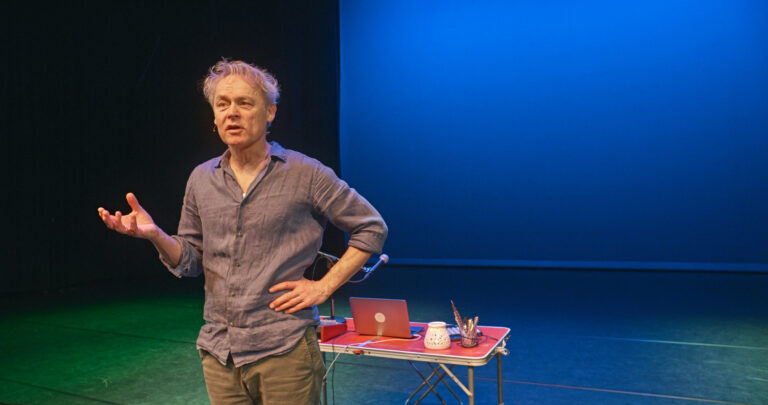 Een man met grijze haren staat met één hand op de heup op een podium, voor een felblauwe achtergrond en een tafel met voorwerpen.