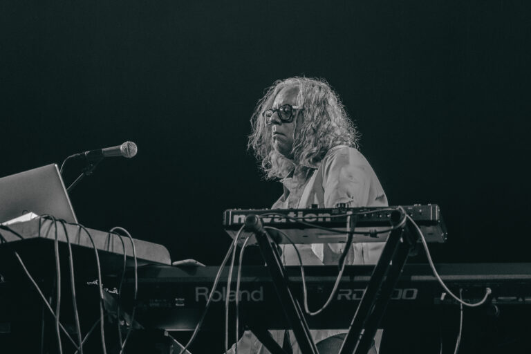 Een muzikant met lange, krullende haren speelt op meerdere synthesizers tijdens een optreden.