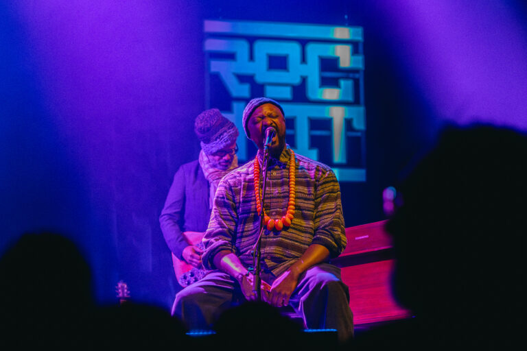 Een muzikant in traditionele kleding zingt op een podium met een blauw paarse lichteffect en een bandlid op de achtergrond.