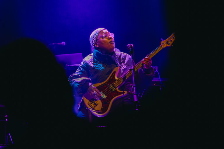 Een muzikant speelt basgitaar op het podium, omringd door paarse verlichting.