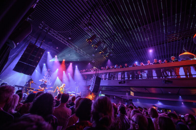 Een druk publiek geniet van een liveoptreden met kleurrijke lichten in een muziekvenue.