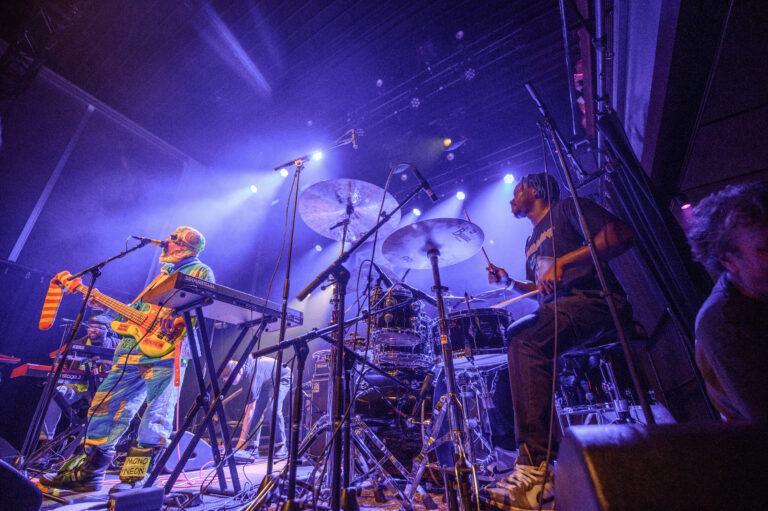 Een muzikant staat op het podium met een gitaar terwijl een drummer en een andere muzikant op de achtergrond spelen, omringd door kleurrijke lichten.