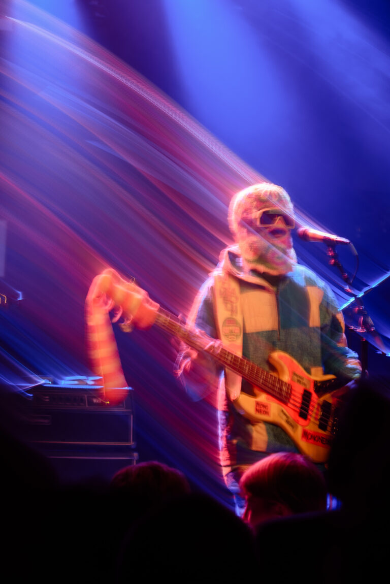 Een muzikant in een kleurrijke outfit speelt gitaar op een podium met sfeervolle lichteffecten.