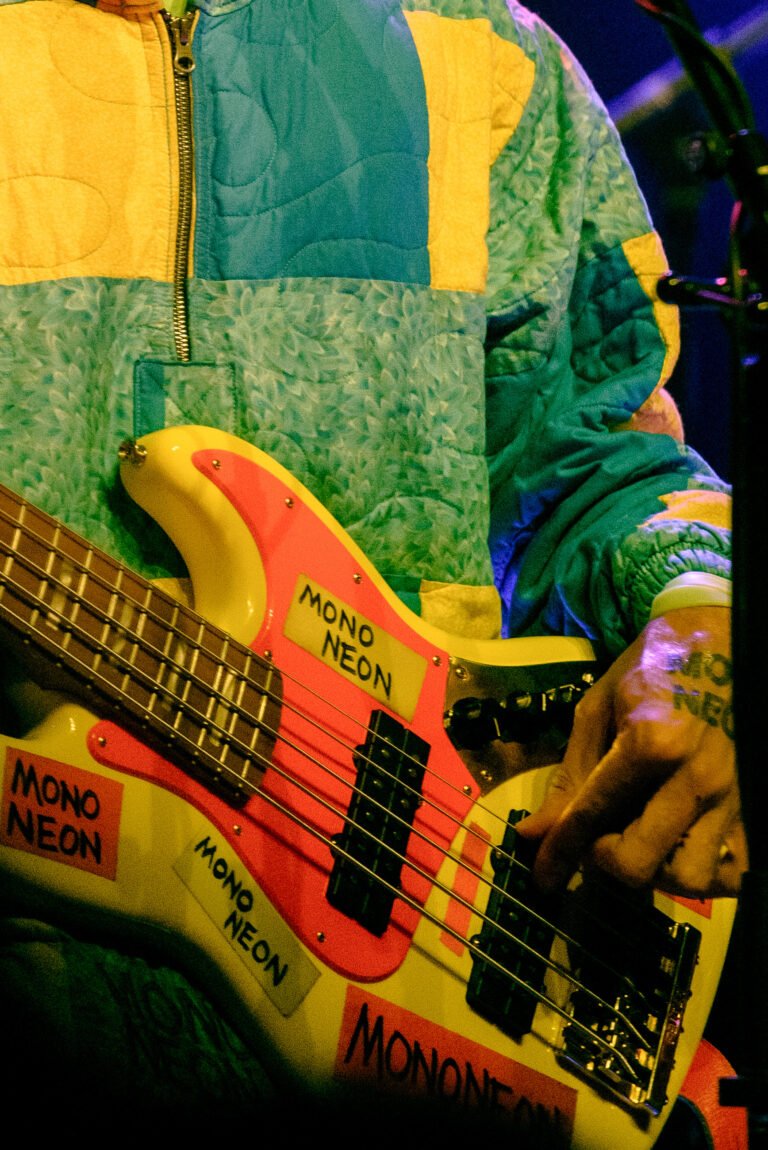 Een close-up van een hand die een opvallende, gekleurde basgitaar bespeelt, met stickers erop.