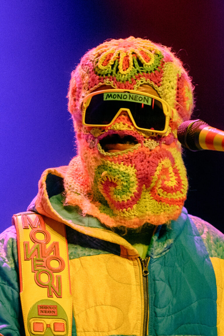 Een persoon in een kleurrijk, zacht gebreid masker en retro zonnebril zingt tijdens een optreden.