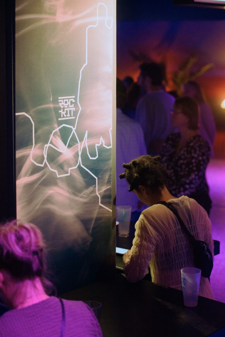 Een vrouw zit aan een bar met haar telefoon, terwijl op de achtergrond mensen in een sfeervol verlichte ruimte mingelen.