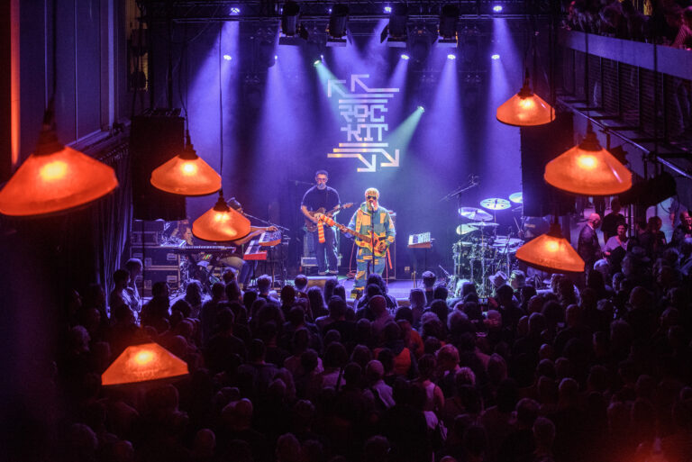 Een muzikant staat op het podium voor een grote menigte, verlicht door paarse lichten en hangende lampen.