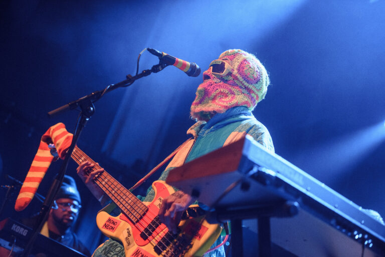 Een muzikant met een kleurrijke hoofdbedekking en zonnebril speelt gitaar op het podium, met een andere muzikant en muziek instrumenten op de achtergrond.