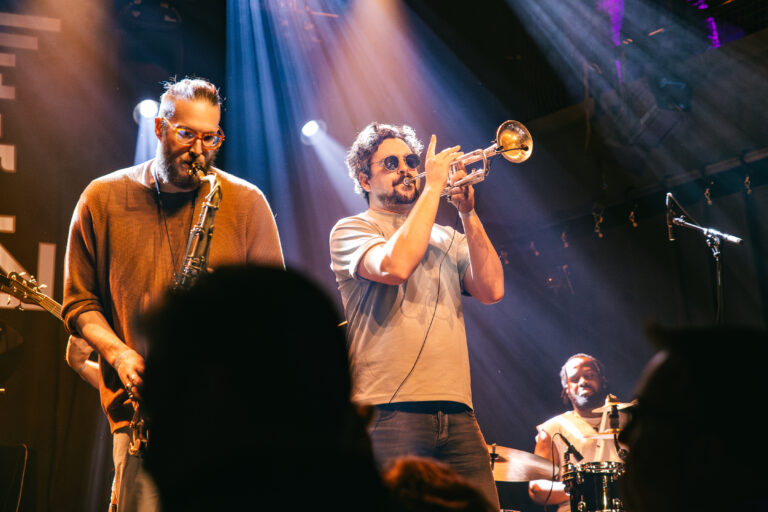 Een band speelt live muziek op het podium met een saxofonist, een trompettist en een drummer, omringd door sfeervolle verlichting.