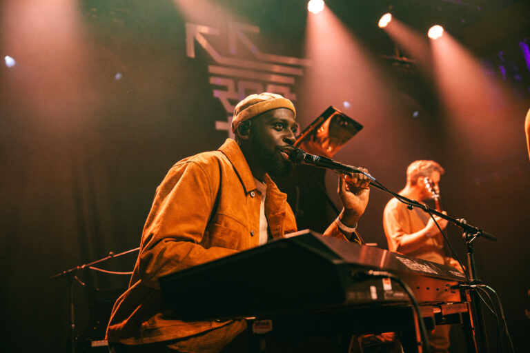 Een muzikant op het podium speelt op een keyboard, terwijl hij in de microfoon zingt, omringd door sfeervolle verlichting.