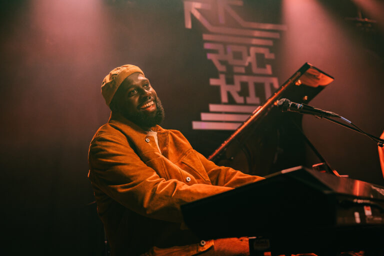 Een muzikant die met een glimlach op een keyboard speelt, omringd door sfeerverlichting.