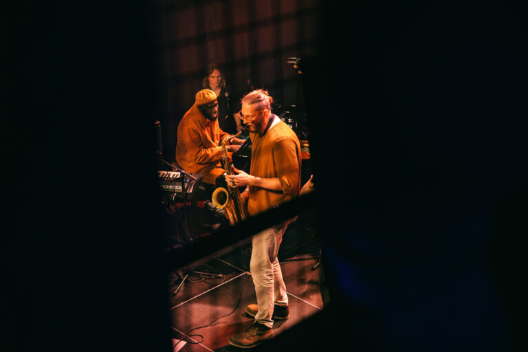 Een muzikant speelt saxofon op een podium, met andere bandleden die in de achtergrond zichtbaar zijn.