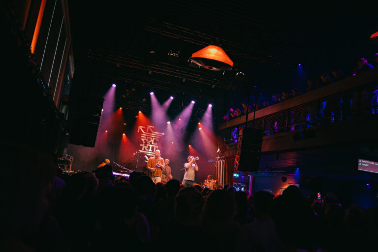 Een live muziekoptreden met een artiest op het podium omringd door kleurrijke verlichting en een enthousiast publiek.
