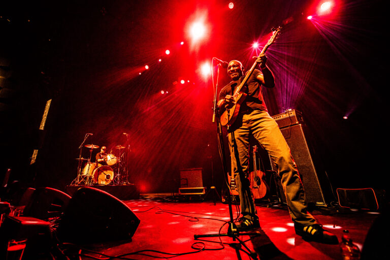 Een muzikant speelt gitaar op een podium met heldere rode lichten en een drumstel op de achtergrond.