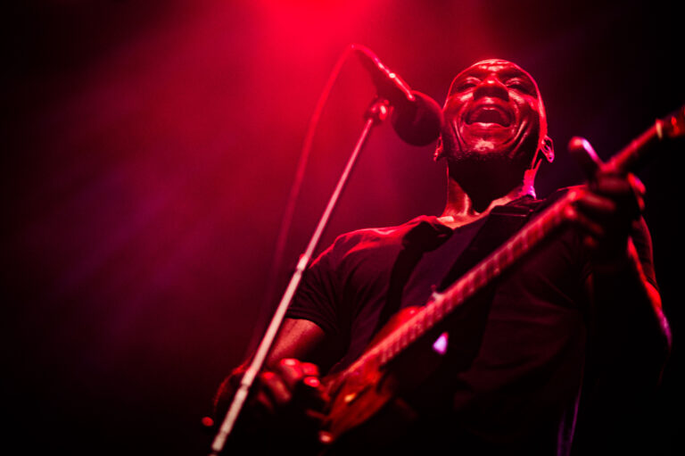 Een muzikant staat op het podium met een microfoon en een snaarinstrument, omringd door rood licht.