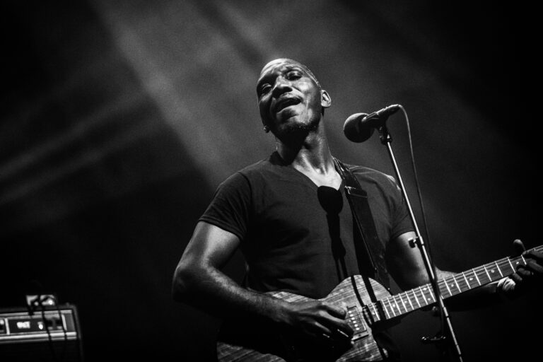 Een muzikant speelt gitaar en zingt op een podium, verlicht door een lichteffect.