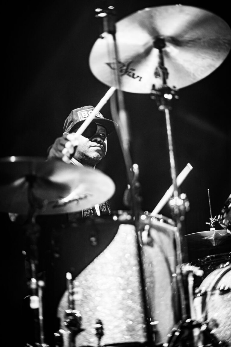Een drummer speelt op een drumstel in een zwart-witfoto, met een focus op de stokken en cymbals.