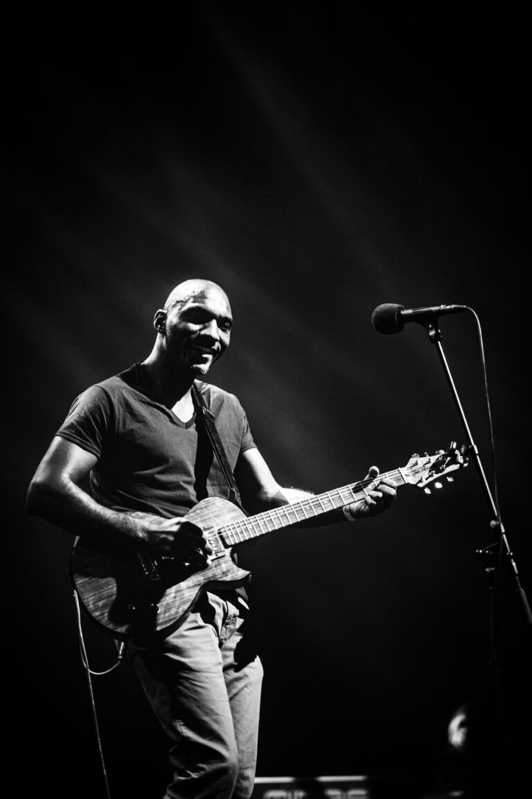 Een muzikant speelt gitaar op een podium in zwart-wit, met een microfoon op de achtergrond.