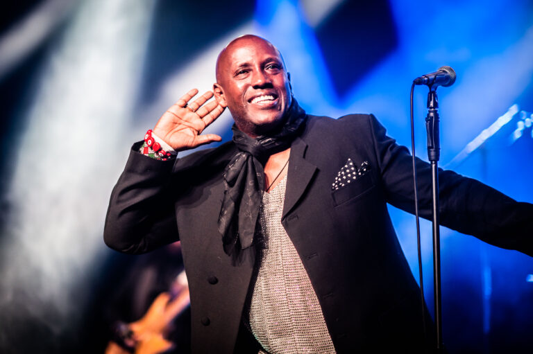 Een glimlachende zanger op het podium die zijn hand achter zijn oor houdt, alsof hij naar het publiek luistert, met kleurrijke verlichting op de achtergrond.