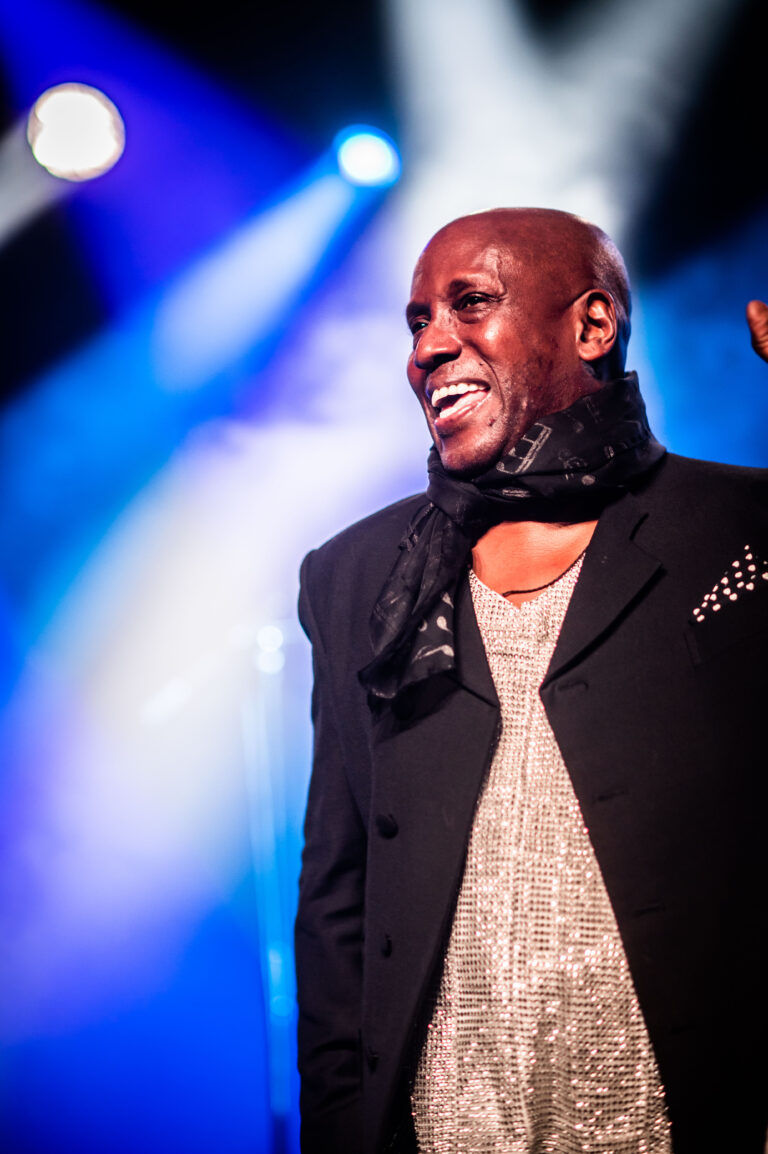 Een lachende man in een zwarte jas en sjaal staat op een podium met gekleurde lichten op de achtergrond.