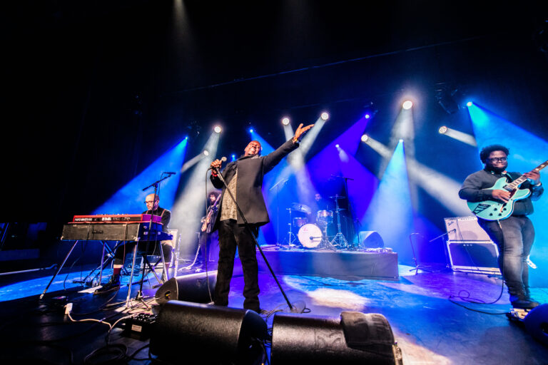 Een muziekgroep treedt op op een podium met kleurrijke lichten, waarbij de zanger een krachtige pose aanneemt.