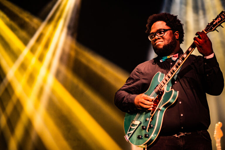 Een muzikant speelt een groene gitaar op een podium met kleurrijke lichtstralen.