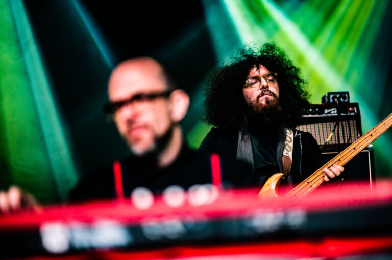 Een muzikant met een grote bos krullend haar speelt basgitaar op het podium, terwijl een andere muzikant op de voorgrond een toetseninstrument bespeelt.