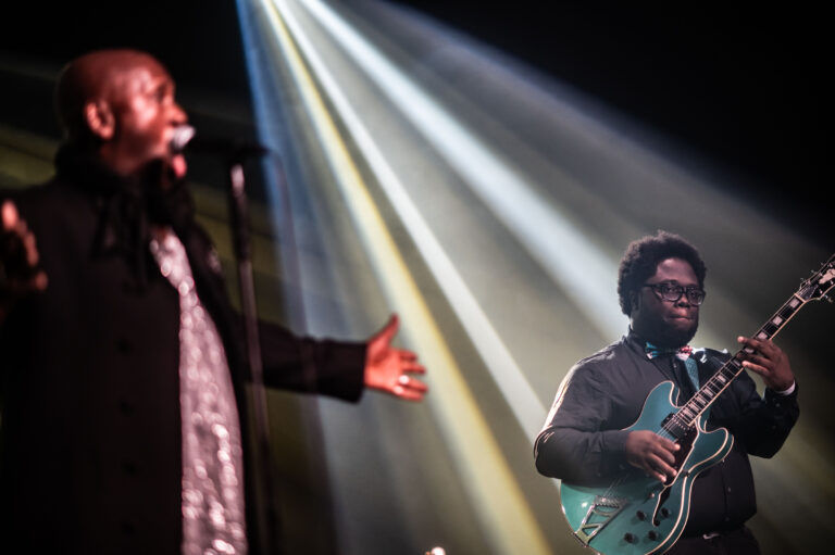 Een zanger staat op het podium met open armen terwijl een gitarist met een lichtblauwe gitaar speelt, omringd door schijnwerpers.