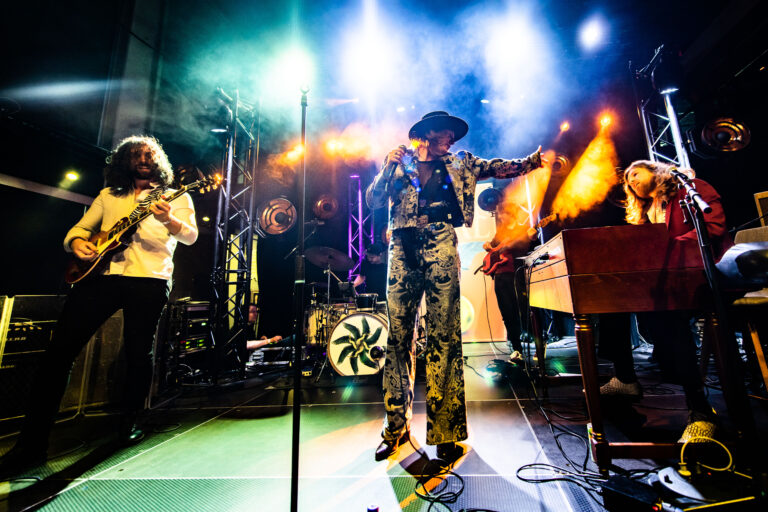 Een liveband die optreedt op een podium, met kleurrijke verlichting en muzikanten die verschillende instrumenten bespelen.