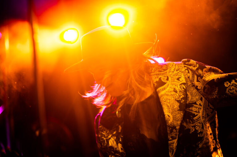 Een silhouet van een muzikant met een hoed, verlicht door fel oranje en paarse lichten op een podium.