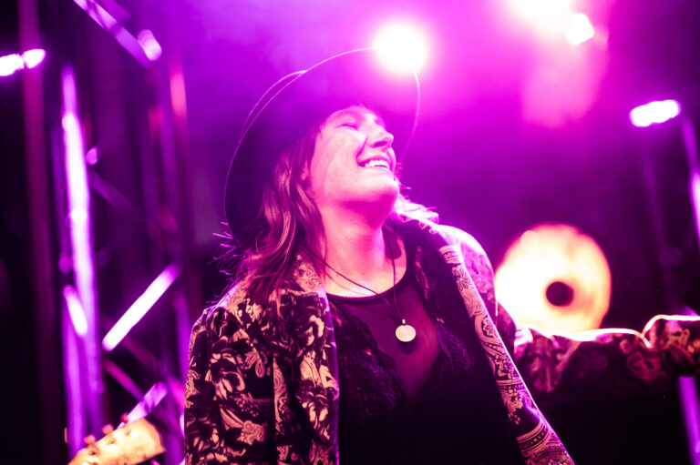 Een zangeres met een hoed glimlacht op het podium, omringd door paarse verlichting.