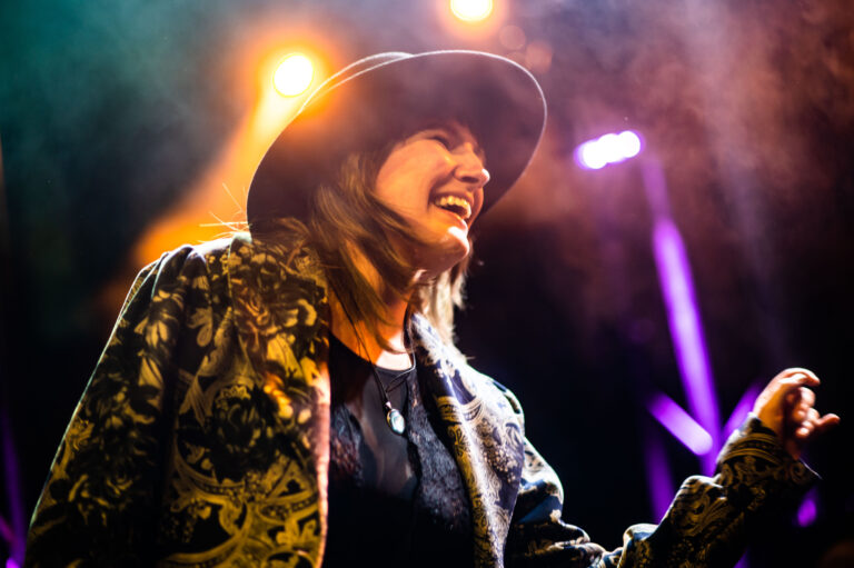 Een vrouw in een hoed zingt enthousiast op het podium, omgeven door kleurrijke lichten.