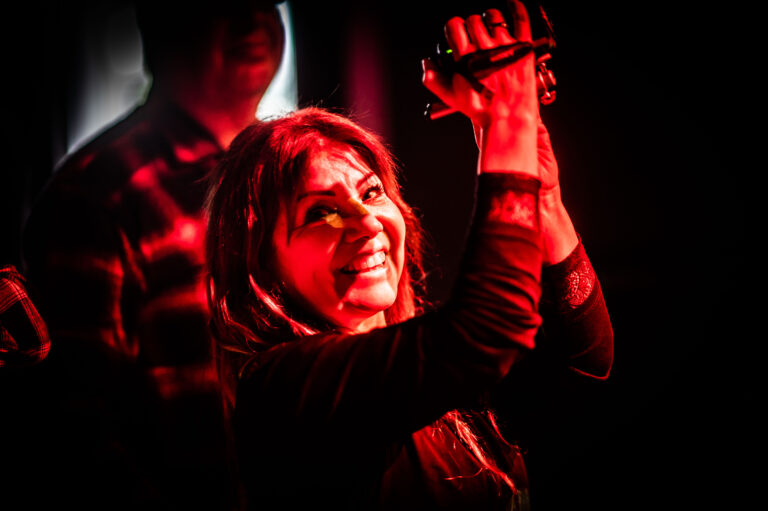 Een vrouw met een glimlach houdt een set maracas vast, omgeven door een rode gloed op het podium.