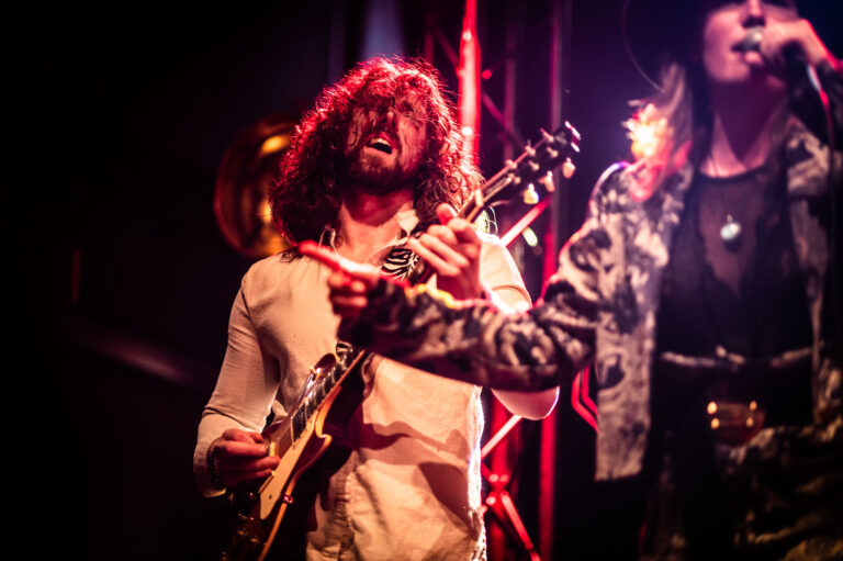 Een muzikant speelt gitaar op een podium, terwijl een andere zangeres in een opvallende outfit zingt.