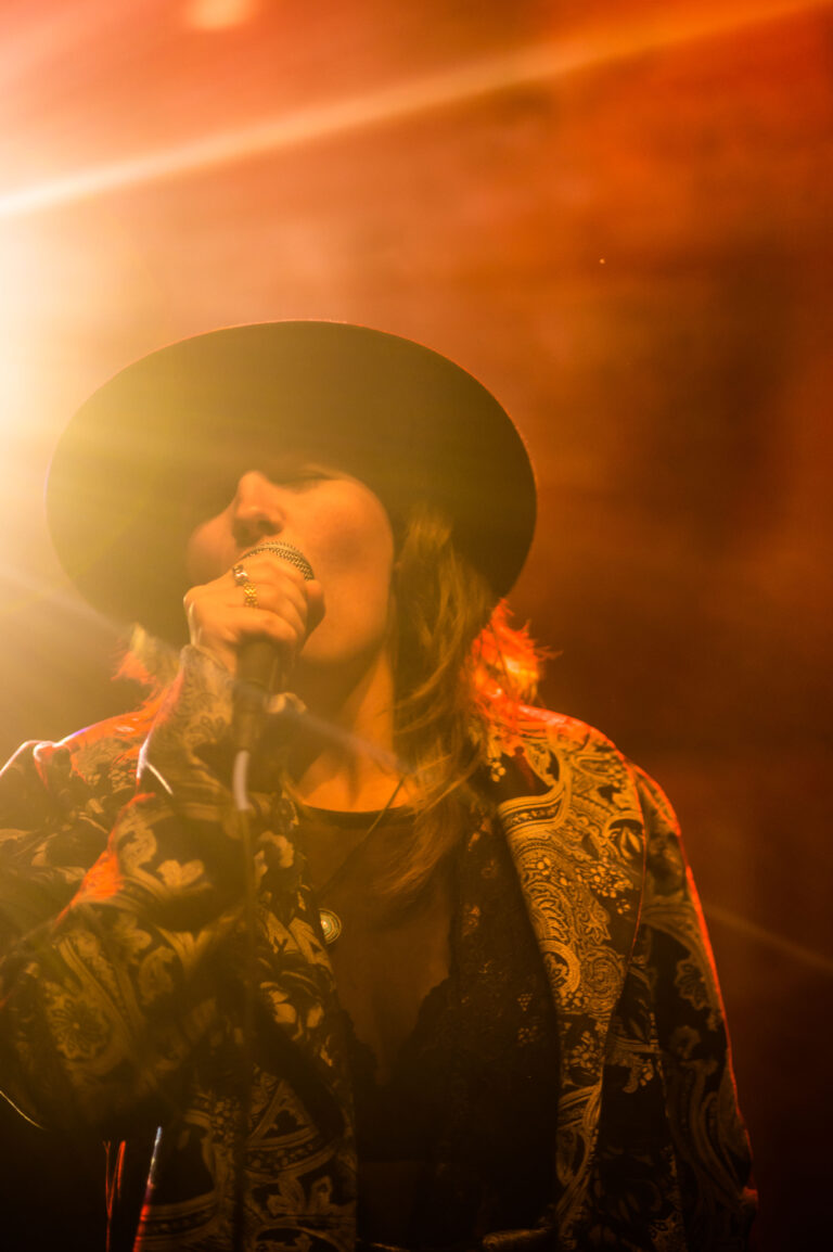 Een zangeres met een hoed staat in een fel verlichte omgeving terwijl ze in een microfoon zingt.