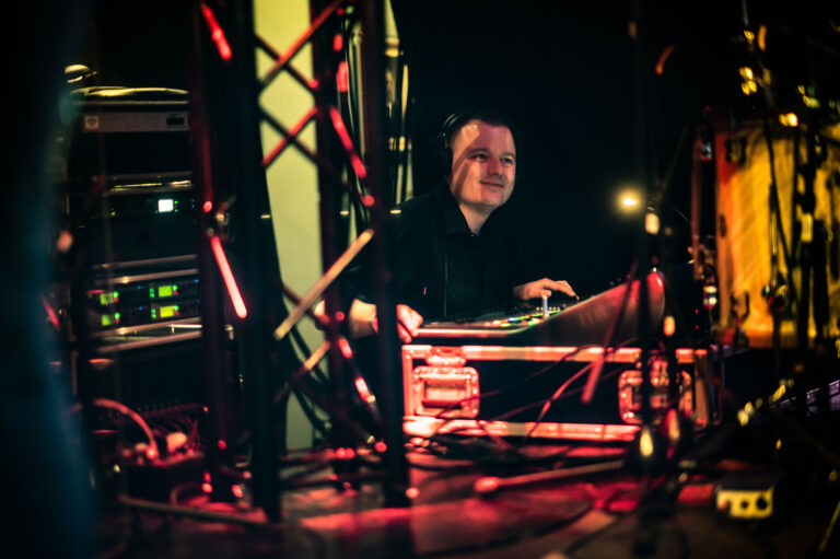 Een technicus zit achter een mengpaneel omringd door kabels en verlichting in een podiumomgeving.