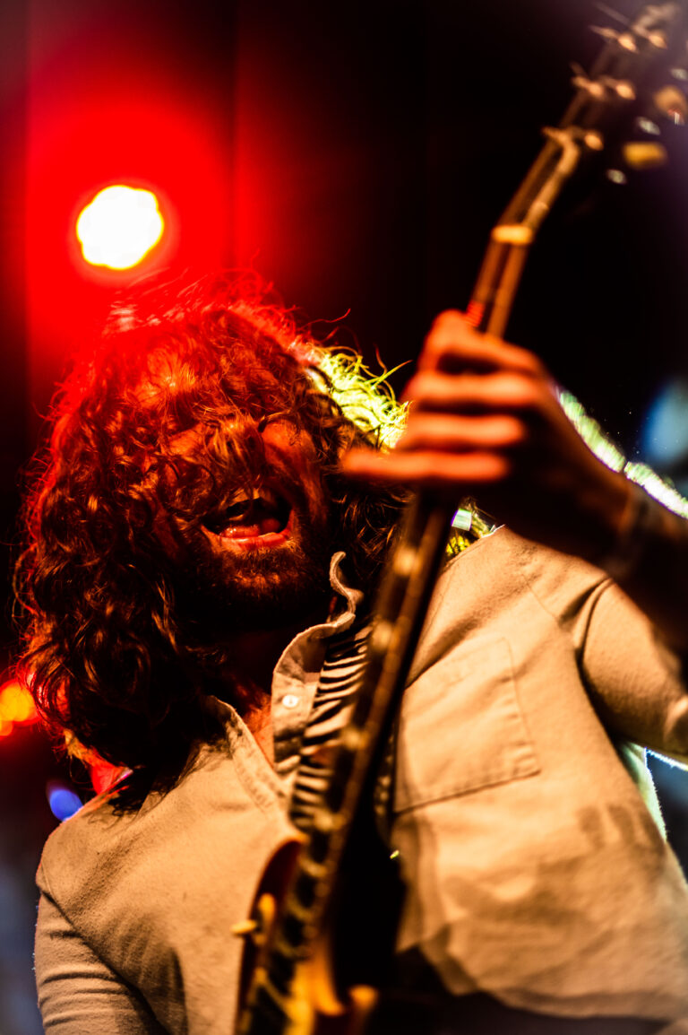Een muzikant met lange, krullende haren speelt op een elektrische gitaar onder heldere lichten.