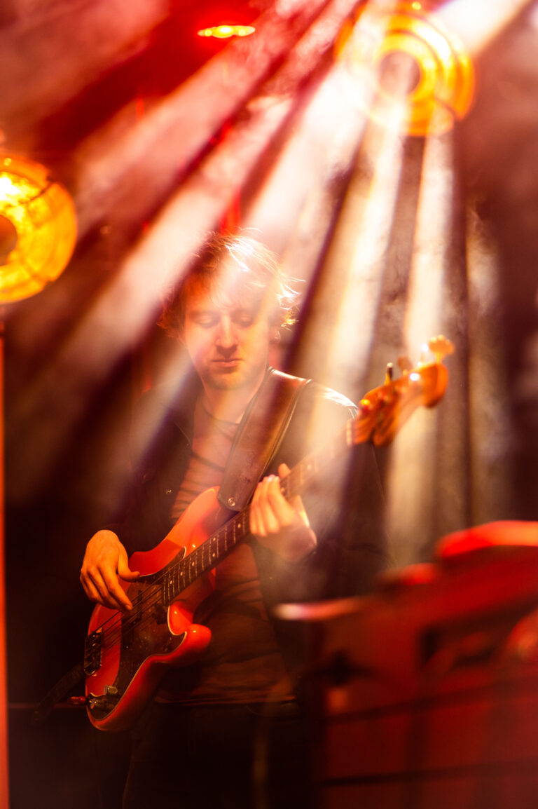 Een muzikant speelt op een elektrische basgitaar, omgeven door lichteffecten en rook in een sfeervolle setting.