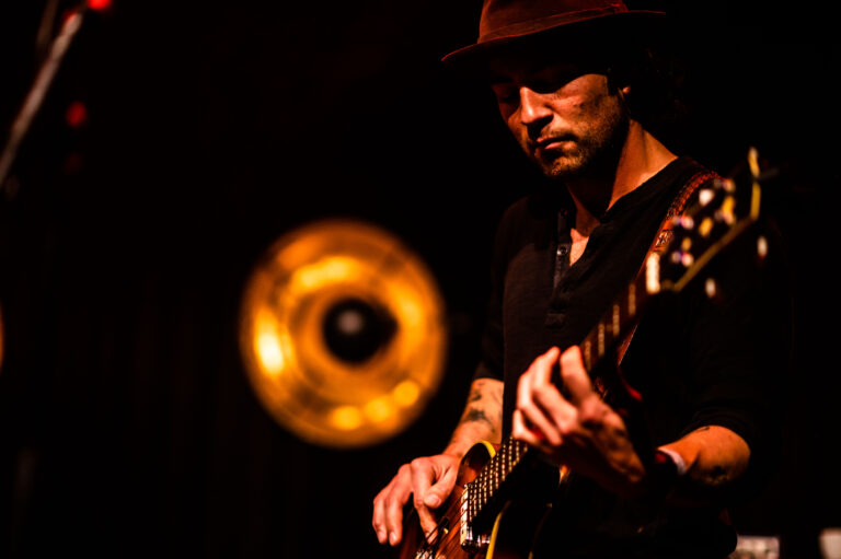 Een muzikant speelt gitaar op het podium, met een vintage trompet op de achtergrond.