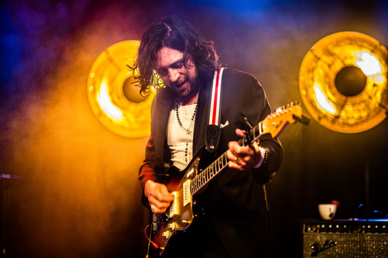 Een muzikant speelt gitaar op het podium, omringd door kleurrijke verlichting en grote luidsprekers.