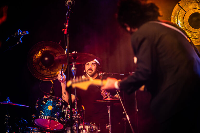 Een drummer in een live muzieksetting speelt energiek op zijn drumstel, terwijl een muzikant op de achtergrond gitaar speelt.