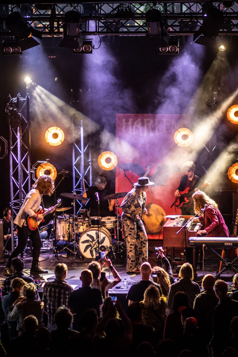 Een band speelt live op een podium met kleurrijke lichten en een enthousiast publiek ervoor.