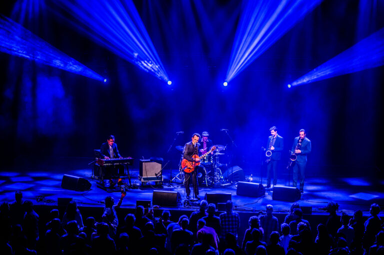 Een band speelt live op het podium, omgeven door een menigte en blauwe schijnwerpers.