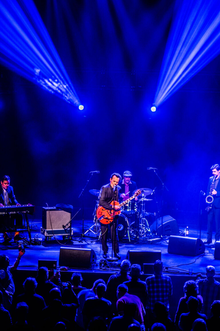 Een band speelt live op een podium met blauwe verlichting en een enthousiast publiek.