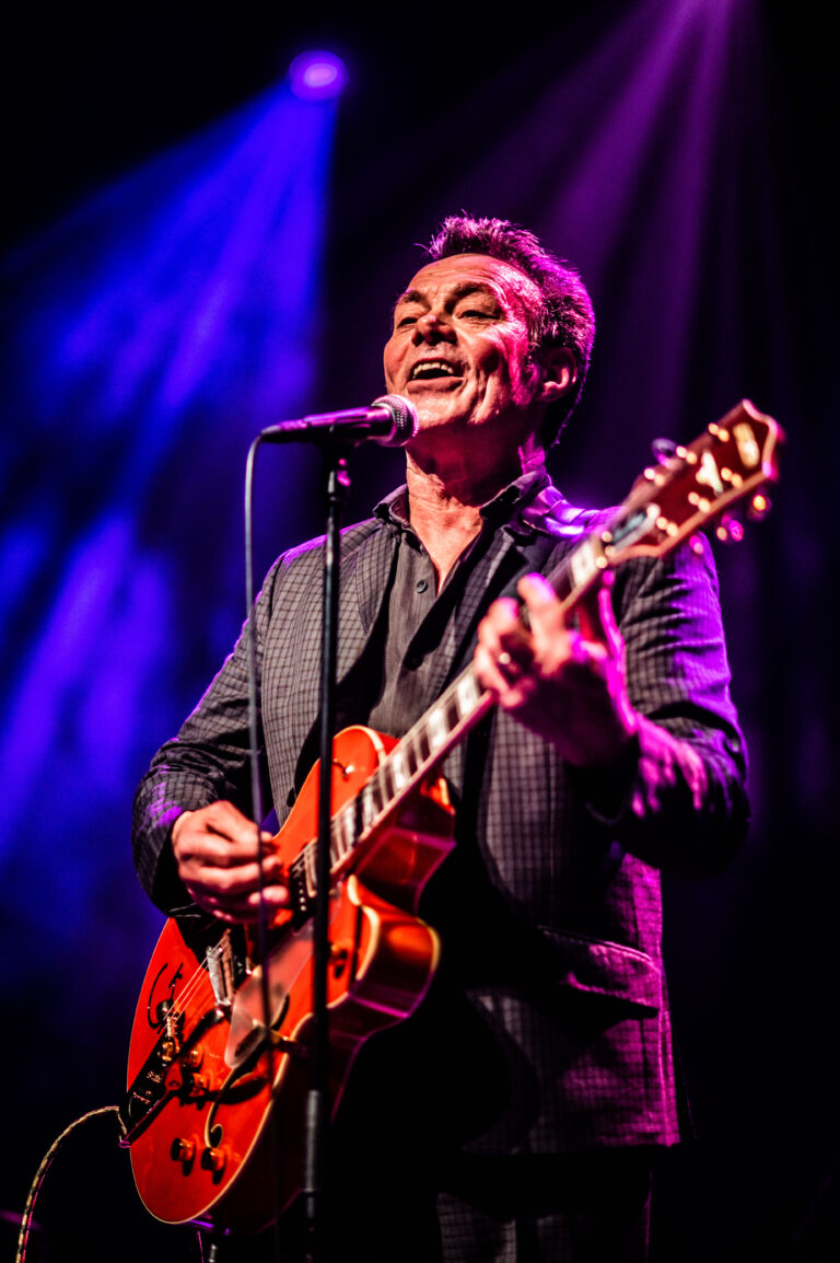 Een muzikant zingt en speelt gitaar op het podium, verlicht door paarse verlichting.