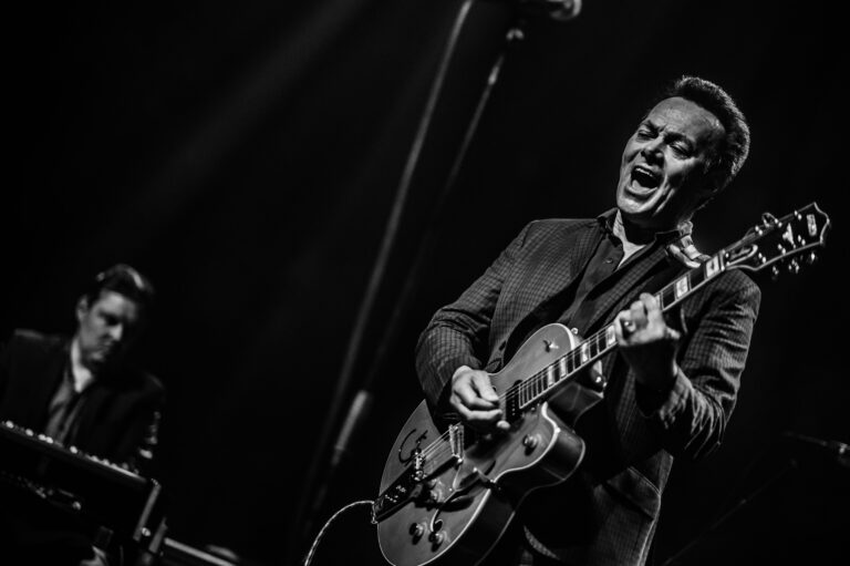 Een muzikant speelt gitaar en zingt op een podium, terwijl een andere muzikant op de achtergrond toetsen speelt.