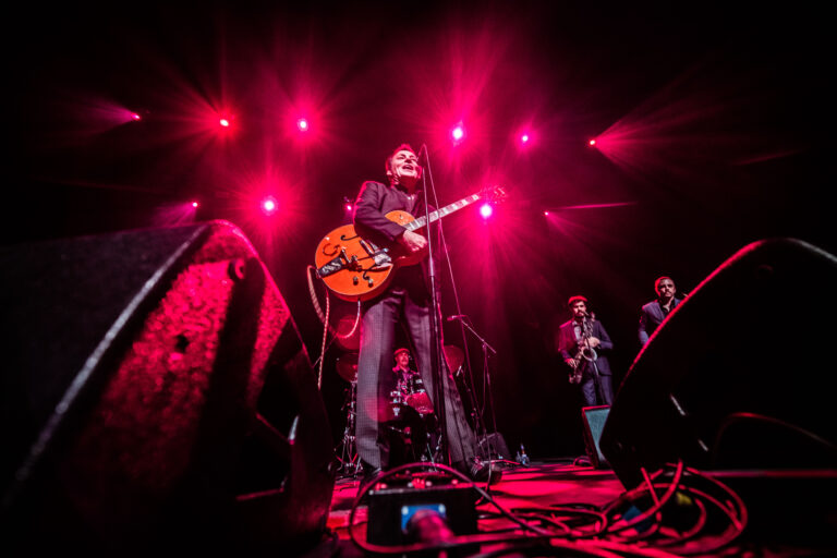 Een muzikant speelt gitaar op een podium, omgeven door roze lichten en bandleden op de achtergrond.