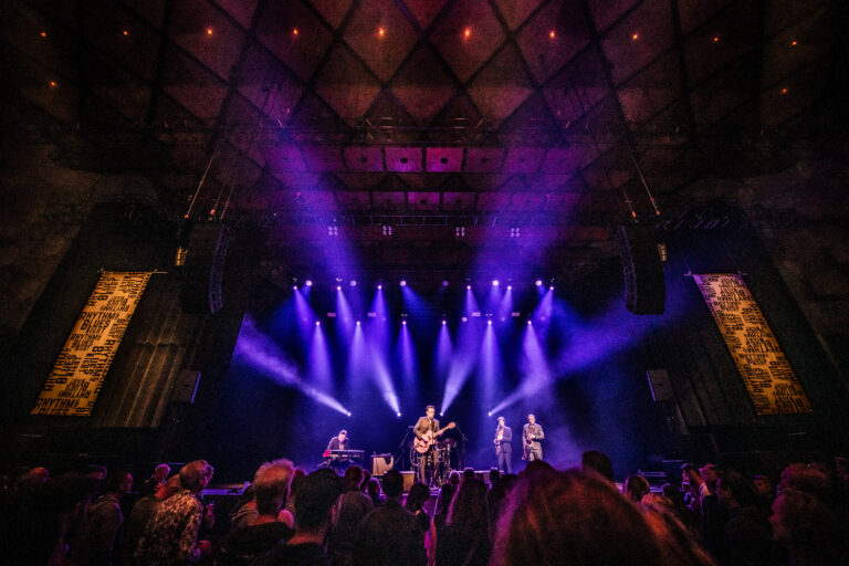 Een muzikale optreden met drie artiesten op een podium, omgeven door een publiek en kleurrijke lichten.